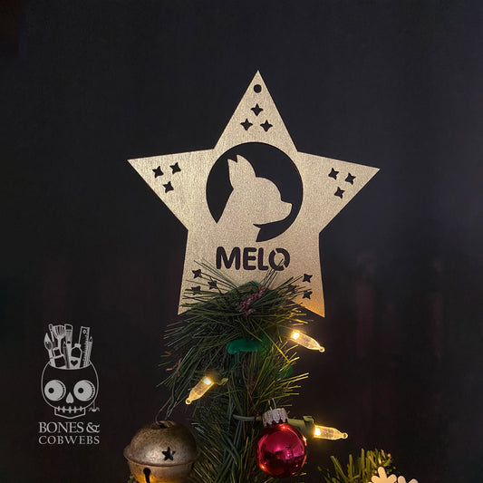 Wooden star-shaped ornament with a pet silhouette and the word 'Melo' cut-out at the bottom, placed on a Christmas tree with lights and decorations.